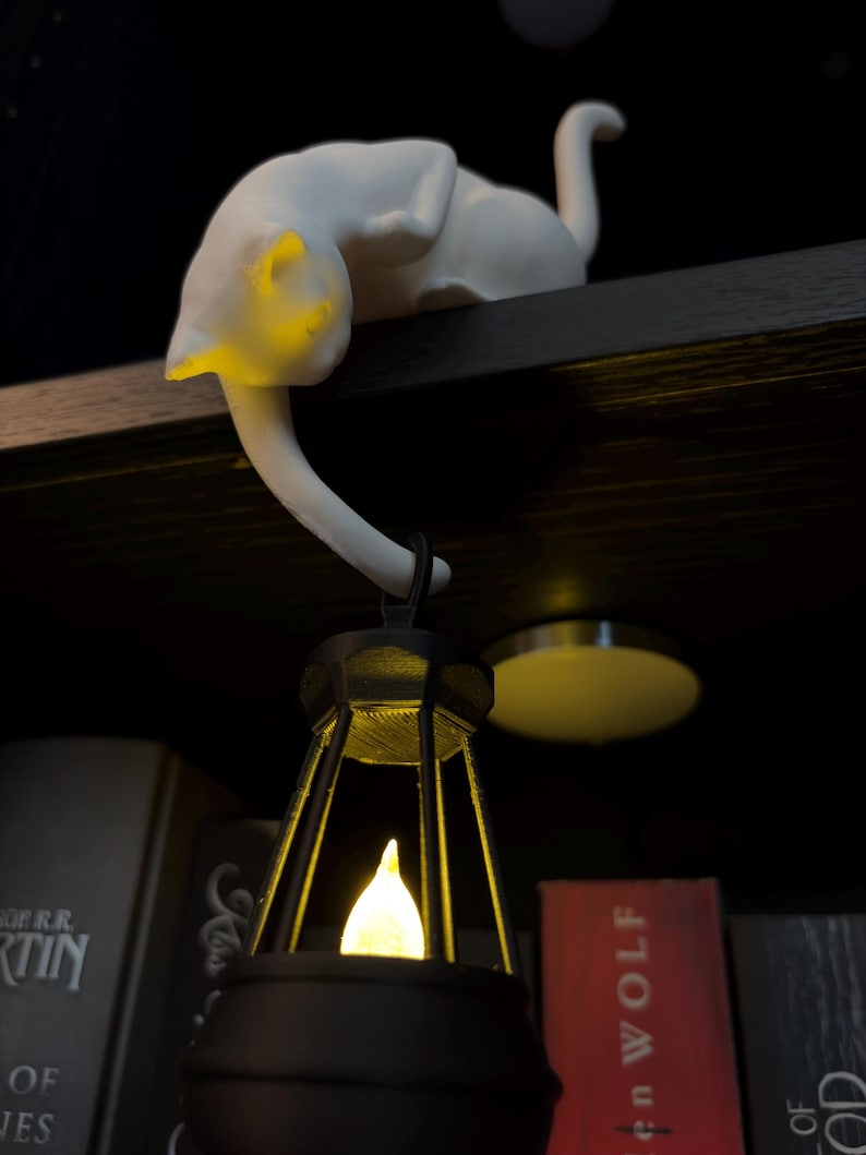 May include: A white cat figurine perched on a ledge, with its tail extending downward to hold a black lantern. The lantern features a glowing yellow light inside, casting a warm illumination on the cat's head and the surrounding area.