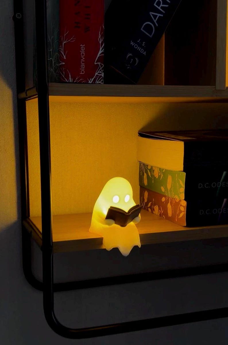 May include: A small, glowing, yellow-tinted ghost-shaped lamp sits on a wooden shelf. The ghost figure has two circular white eyes and is holding a small, dark, open book in its hands. The lamp emits a warm, soft light that illuminates the surrounding area.