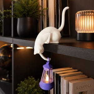 May include: A white cat figurine in a crouching position perched on a shelf edge, with its tail hanging down to hold a small purple lantern. The lantern features a glowing, warm-toned LED light inside, creating a soft illumination effect.