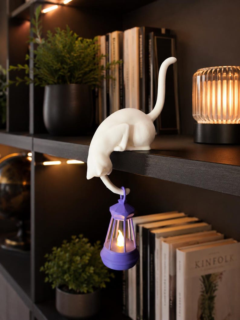 May include: A white cat figurine in a crouching position perched on a shelf edge, with its tail hanging down to hold a small purple lantern. The lantern features a glowing, warm-toned LED light inside, creating a soft illumination effect.
