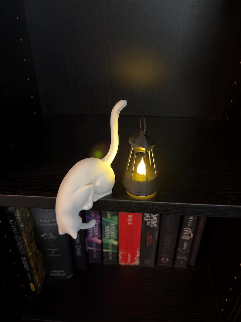 May include: A white cat-shaped lamp with an upright tail and a small, dark-colored lantern emitting a warm yellow glow. Both items are positioned on a shelf above a row of books. The cat lamp features a smooth, matte finish and a curved posture.