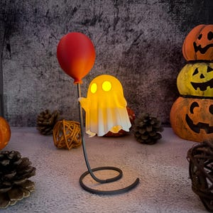 May include: A Halloween ghost lamp with a red balloon. The ghost, lit with a warm yellow glow, is draped in a white sheet. The lamp has a black, swirled base. Pumpkins and pinecones are in the background, creating a spooky atmosphere.