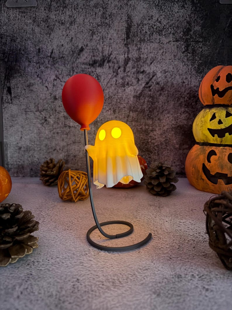May include: A Halloween ghost lamp with a red balloon. The ghost, lit with a warm yellow glow, is draped in a white sheet. The lamp has a black, swirled base. Pumpkins and pinecones are in the background, creating a spooky atmosphere.