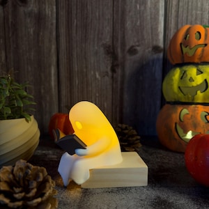 May include: A small, glowing white ghost figurine sitting on a light-colored wooden block, holding and reading a miniature black book. The ghost emits a warm, soft yellow light from within its translucent body, creating a gentle illumination.