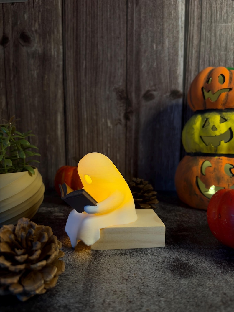 May include: A small, glowing white ghost figurine sitting on a light-colored wooden block, holding and reading a miniature black book. The ghost emits a warm, soft yellow light from within its translucent body, creating a gentle illumination.