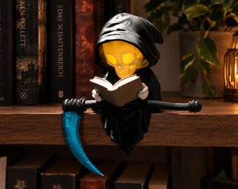 May include: A small figurine of a hooded skeleton reading an open book, featuring a glowing yellow skull and a bright blue scythe blade. The figure is designed to sit on the edge of a shelf, with the scythe handle extending horizontally.