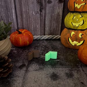 May include: Two small, open book-shaped charms are positioned side-by-side. One charm is a solid matte brown color, while the other is a vibrant, light-emitting neon green. Both items feature a simple, minimalist design with visible pages.