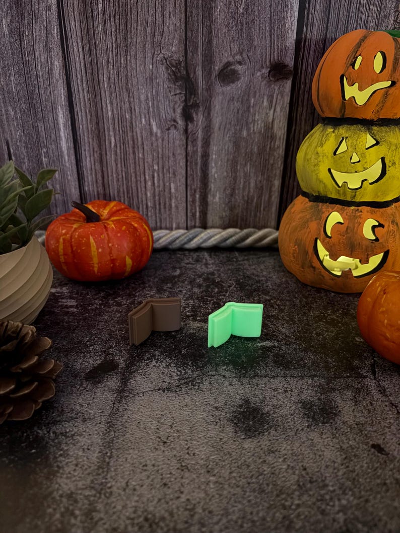 May include: Two small, open book-shaped charms are positioned side-by-side. One charm is a solid matte brown color, while the other is a vibrant, light-emitting neon green. Both items feature a simple, minimalist design with visible pages.