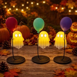 May include: Three Halloween ghost lights with balloons. Each yellow ghost has glowing eyes and a white bottom, held by a black stand. A red, green, and purple balloon floats above each ghost. Pumpkins and autumn leaves are in the background.