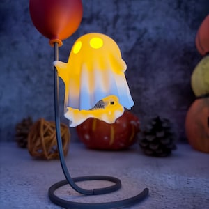 May include: A Halloween-themed lamp with a glowing yellow ghost holding a red balloon. The ghost has three yellow eye-shaped lights. The lamp has a dark grey base and stem. Pumpkins and pinecones are in the background, creating a festive atmosphere.