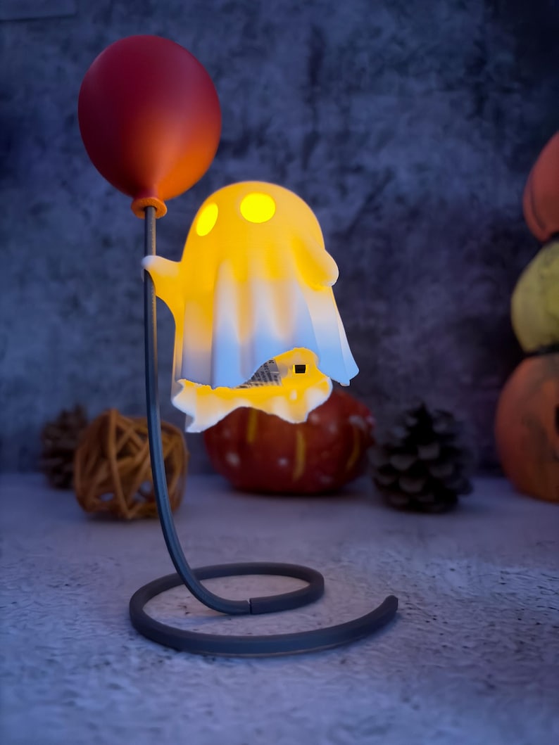 May include: A Halloween-themed lamp with a glowing yellow ghost holding a red balloon. The ghost has three yellow eye-shaped lights. The lamp has a dark grey base and stem. Pumpkins and pinecones are in the background, creating a festive atmosphere.