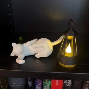 May include: A white cat figurine lying on its side next to a black lantern with a glowing yellow light inside. The cat is positioned horizontally with its tail extended, while the lantern features a handle at the top and a flame-shaped bulb.