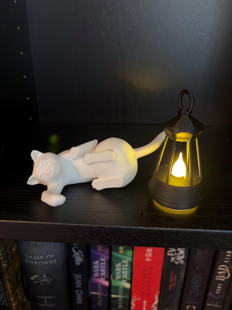 May include: A white cat figurine lying on its side next to a black lantern with a glowing yellow light inside. The cat is positioned horizontally with its tail extended, while the lantern features a handle at the top and a flame-shaped bulb.