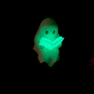 May include: A small, glowing, light-green figurine shaped like a ghost holding an open book. The ghost has two dark, circular eye cutouts and a smooth, rounded head. The entire figure emits a bright, neon-green light against a black background.