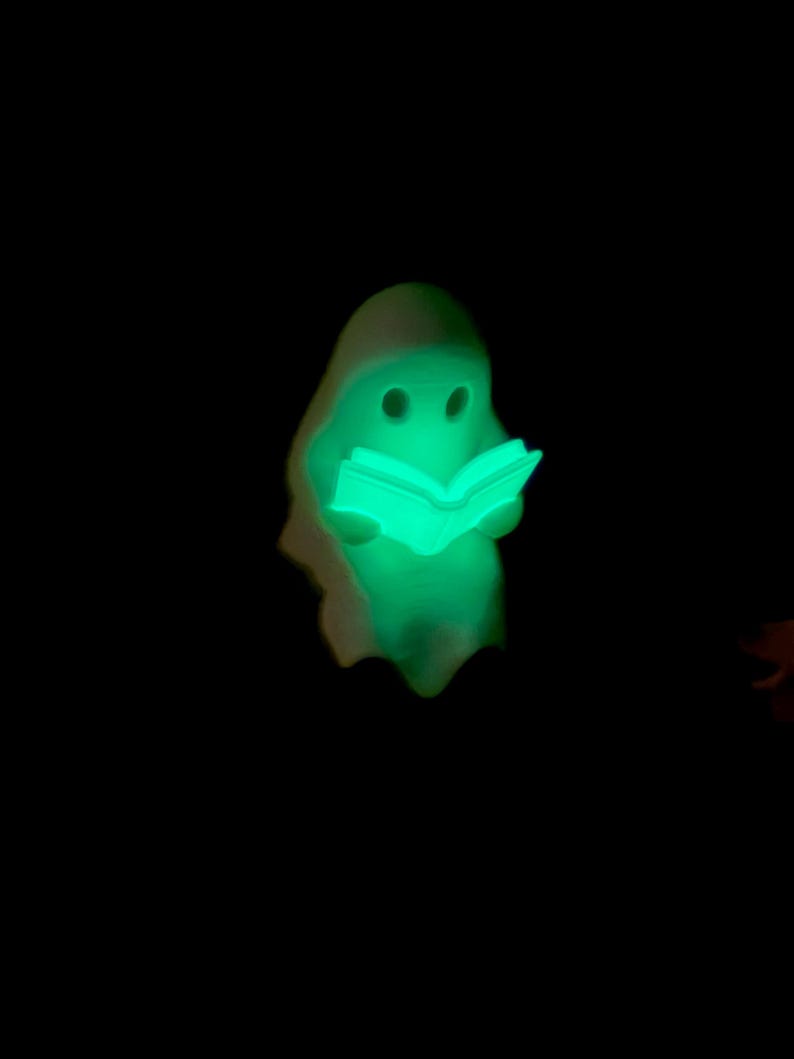 May include: A small, glowing, light-green figurine shaped like a ghost holding an open book. The ghost has two dark, circular eye cutouts and a smooth, rounded head. The entire figure emits a bright, neon-green light against a black background.