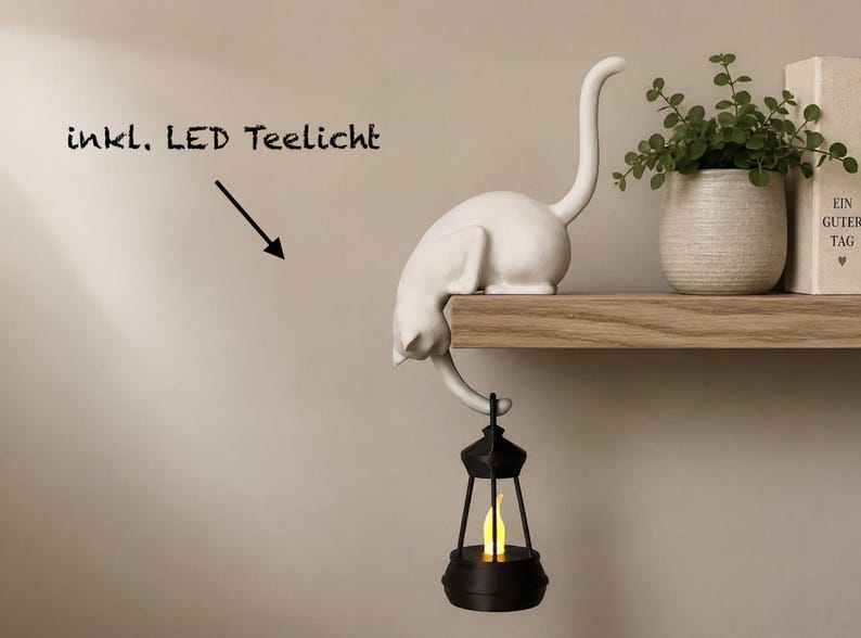 May include: A white cat figurine perched on the edge of a shelf with its tail hanging down, holding a black lantern that contains a glowing yellow LED tea light. The text "inkl. LED Teelicht" is visible with an arrow pointing to the lantern.