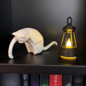 May include: A white cat figurine in a seated, grooming posture positioned next to a small, black, lantern-style light fixture that emits a warm, glowing yellow light from its center, both resting on a dark shelf above a row of books.