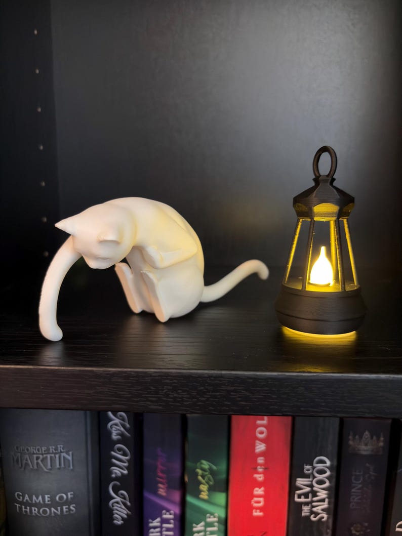 May include: A white cat figurine in a seated, grooming posture positioned next to a small, black, lantern-style light fixture that emits a warm, glowing yellow light from its center, both resting on a dark shelf above a row of books.
