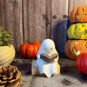 May include: A small white ghost figurine with hollow eyes sitting on a light-colored wooden block while holding a brown open book. The ghost has a smooth, matte finish and is positioned in a central upright posture, displaying a simple form.