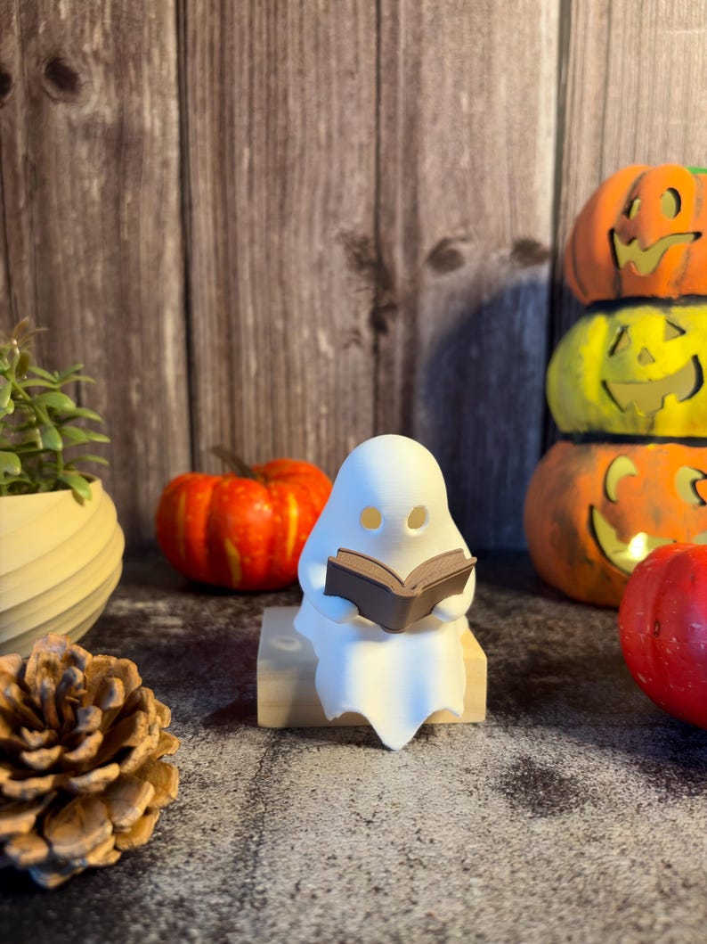 May include: A small white ghost figurine with hollow eyes sitting on a light-colored wooden block while holding a brown open book. The ghost has a smooth, matte finish and is positioned in a central upright posture, displaying a simple form.