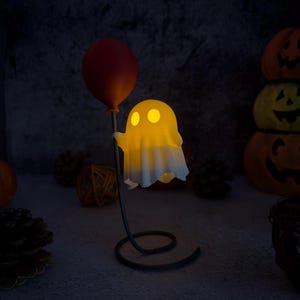 May include: A lit-up ghost ornament holding a red balloon. The ghost has two yellow eyes and a white sheet-like body. The figurine is on a black metal stand with a spiral base. Pumpkins and pinecones are in the background.