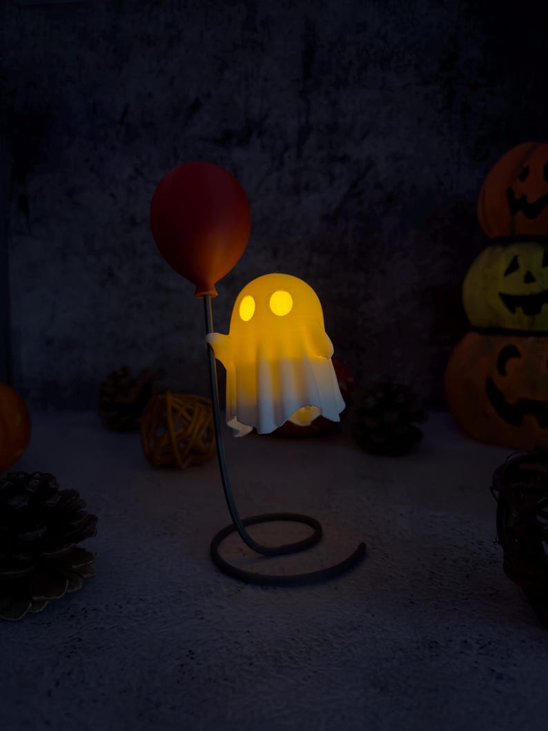 May include: A lit-up ghost ornament holding a red balloon. The ghost has two yellow eyes and a white sheet-like body. The figurine is on a black metal stand with a spiral base. Pumpkins and pinecones are in the background.