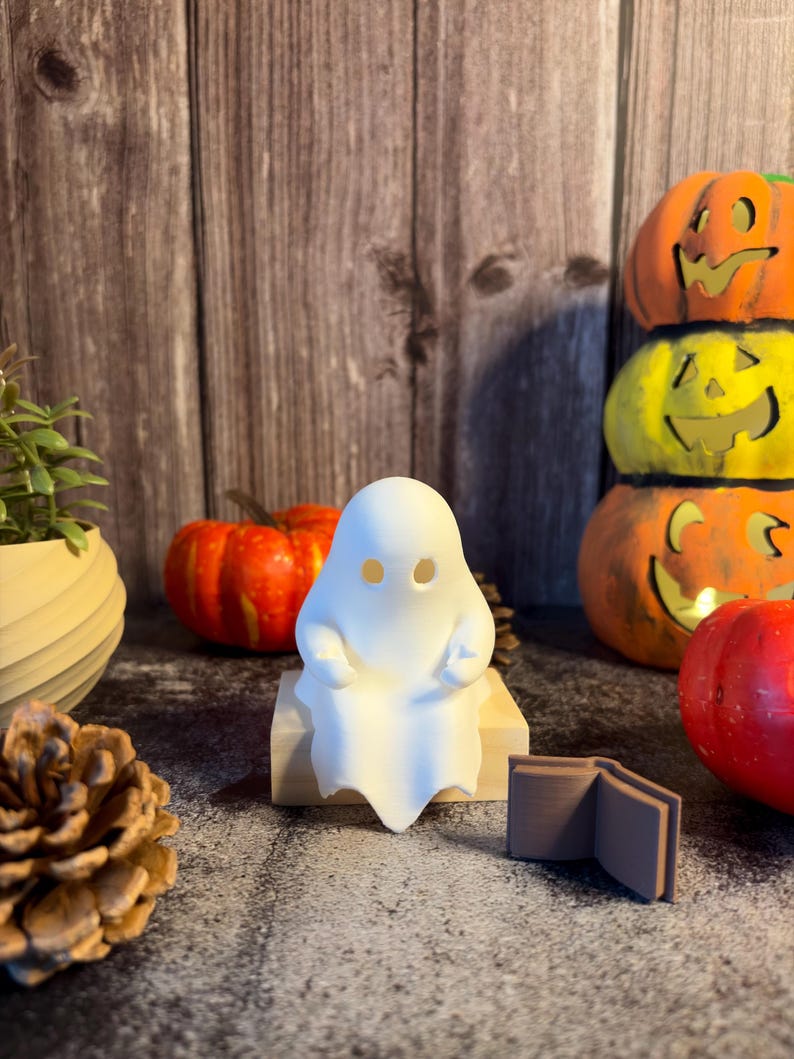 May include: A small, matte white 3D-printed ghost figurine with two circular eye cutouts, sitting on a light-colored wooden block. Beside the ghost is a small, brown, open book figurine, positioned on a dark, textured surface.