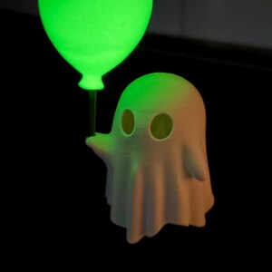 May include: A 3D-printed ghost figure with a glowing green balloon. The ghost is white with eye holes and a draped form. The balloon emits a bright green light, contrasting against the dark background. A fun, spooky design.
