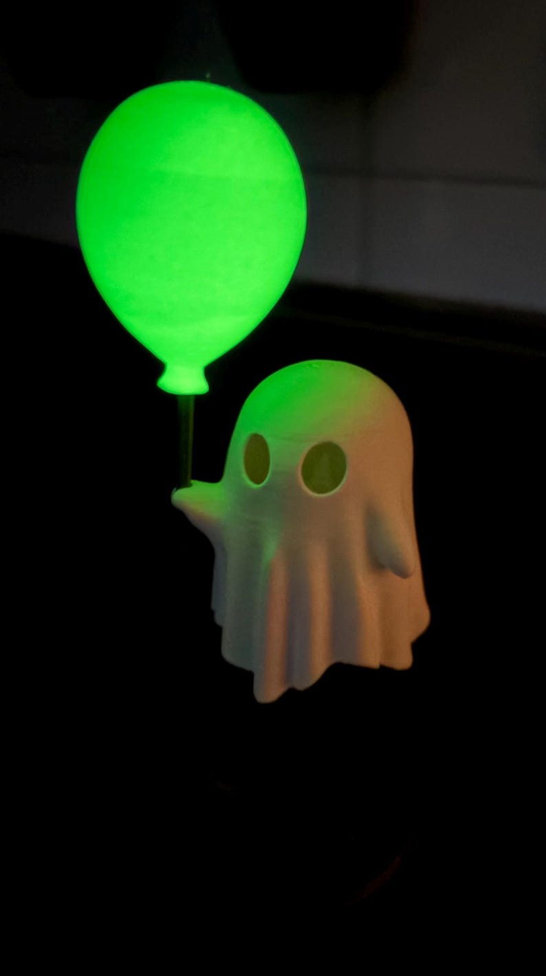 May include: A 3D-printed ghost figure with a glowing green balloon. The ghost is white with eye holes and a draped form. The balloon emits a bright green light, contrasting against the dark background. A fun, spooky design.