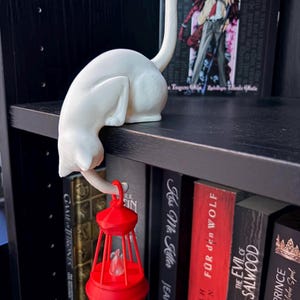 May include: A white cat figurine in a crouching position with its tail extended upward, holding a small, bright red lantern with a clear flame-shaped insert. The cat is perched on the edge of a black shelf, positioned in front of several books.