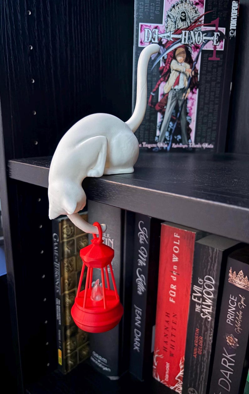 May include: A white cat figurine in a crouching position with its tail extended upward, holding a small, bright red lantern with a clear flame-shaped insert. The cat is perched on the edge of a black shelf, positioned in front of several books.