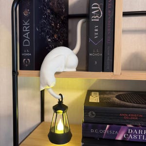 May include: A white cat figurine perched on a shelf edge, featuring a long tail that curves downward to hold a small black lantern. The lantern contains a glowing yellow light, illuminating the space below the cat figure on the wooden shelf.