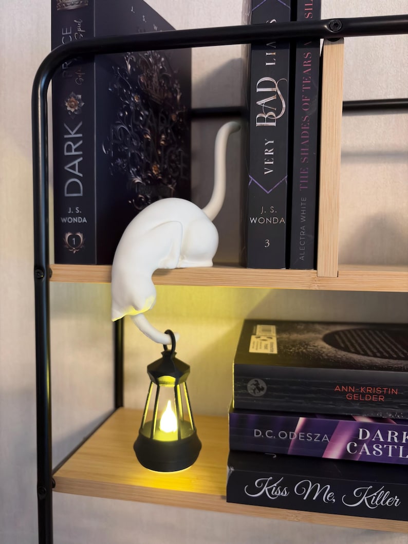 May include: A white cat figurine perched on a shelf edge, featuring a long tail that curves downward to hold a small black lantern. The lantern contains a glowing yellow light, illuminating the space below the cat figure on the wooden shelf.