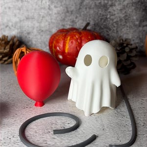 May include: A Halloween display with a red balloon, a white ghost figure, and a grey curved object. An orange pumpkin is in the background. The items are arranged on a textured grey surface.