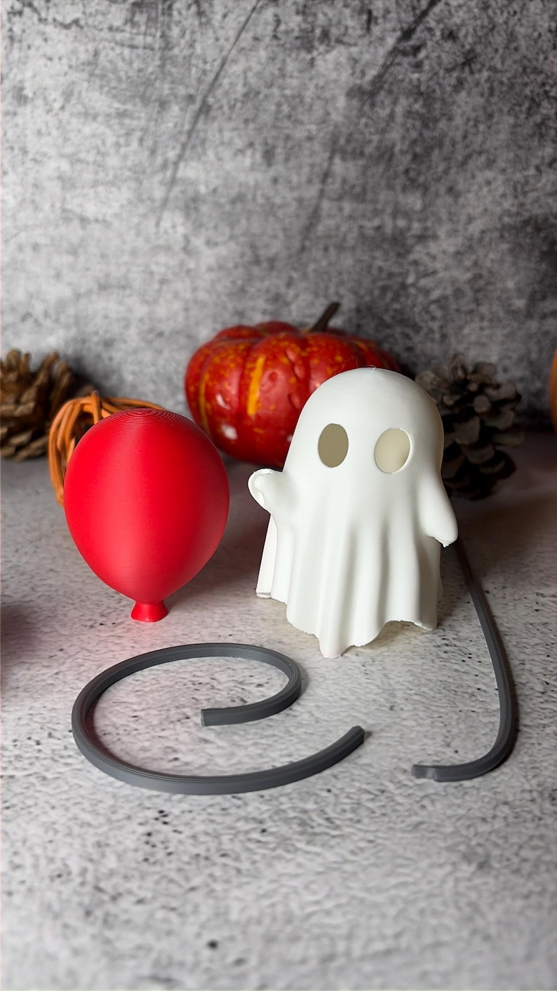 May include: A Halloween display with a red balloon, a white ghost figure, and a grey curved object. An orange pumpkin is in the background. The items are arranged on a textured grey surface.