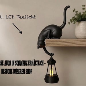 May include: A black cat figurine perched on the edge of a shelf, with its tail extending downward to hold a small black lantern containing a glowing yellow LED tea light. The text "inkl. LED Teelicht" and "WAHLWEISE AUCH IN SCHWARZ ERH&Auml;LTLICH - BESUCHE UNSEREN SHOP" is visible.