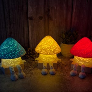 Könnte beinhalten: Drei pilzförmige Lampen mit farbigen, perforierten Kappen und leuchtenden Körpern. Die Lampen haben Beine und stehen auf Holzsockeln. Die Kappen sind türkis, gelb und rot. Die Lampen sind beleuchtet und erzeugen eine warme Atmosphäre.