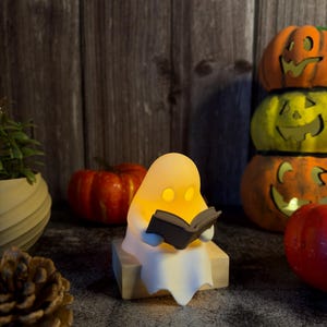 May include: A small, white, glowing ghost figurine sitting on a light-colored wooden block, holding an open dark gray book. The ghost has two circular cutouts for eyes, emitting a warm yellow light from within its smooth, rounded body.