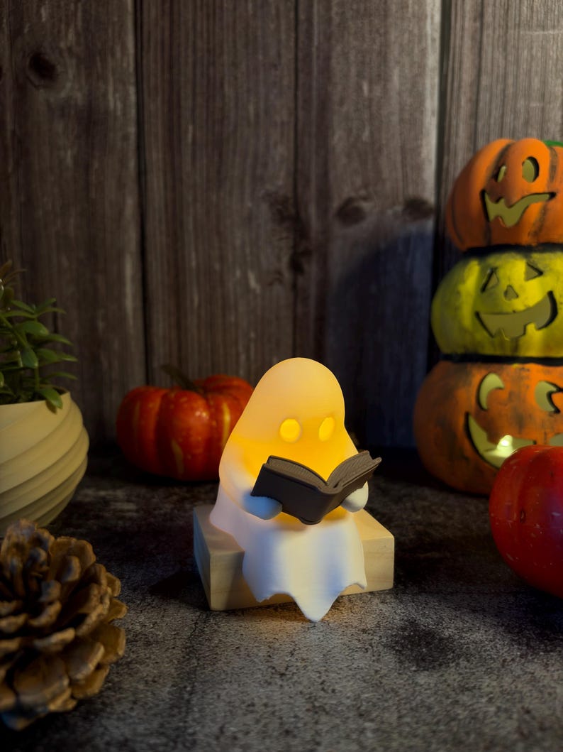 May include: A small, white, glowing ghost figurine sitting on a light-colored wooden block, holding an open dark gray book. The ghost has two circular cutouts for eyes, emitting a warm yellow light from within its smooth, rounded body.