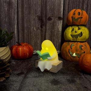 May include: A small, glowing yellow ghost figurine sitting on a light-colored wooden block, holding an open light green book. The ghost has a smooth, rounded shape and emits a soft light from within, creating a warm, illuminated appearance.