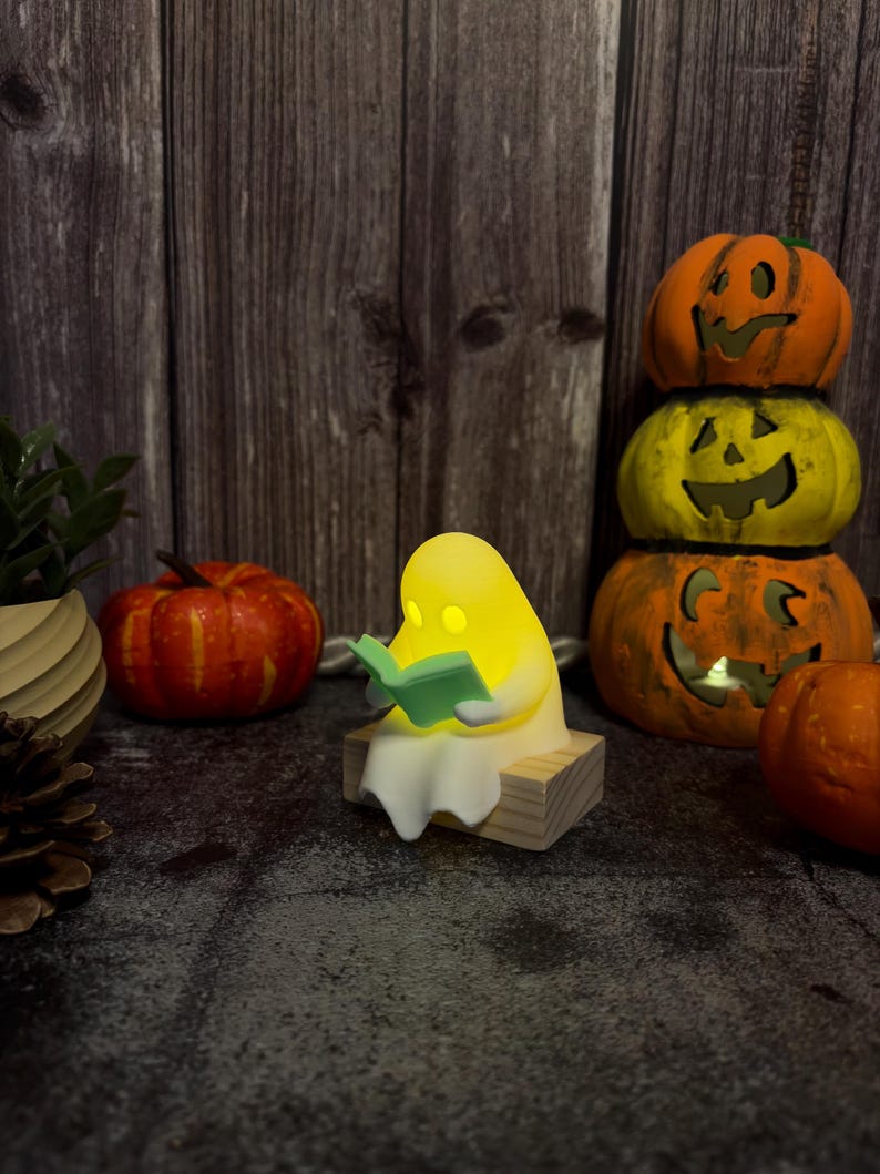 May include: A small, glowing yellow ghost figurine sitting on a light-colored wooden block, holding an open light green book. The ghost has a smooth, rounded shape and emits a soft light from within, creating a warm, illuminated appearance.