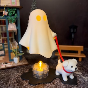 Könnte beinhalten: Eine skurrile Halloween-Figur, die einen weißen Geist mit gelben Augen zeigt, der eine rote Leine hält, die mit einem kleinen weißen Hund verbunden ist. Eine leuchtende Kerze steht auf einem schwarzen Sockel und erzeugt eine gruselige Atmosphäre.