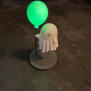 May include: A Halloween-themed ornament depicting a white ghost figure holding a glowing green balloon. The ghost has eye holes and a draped sheet design. The balloon and ghost are mounted on a dark grey circular base.
