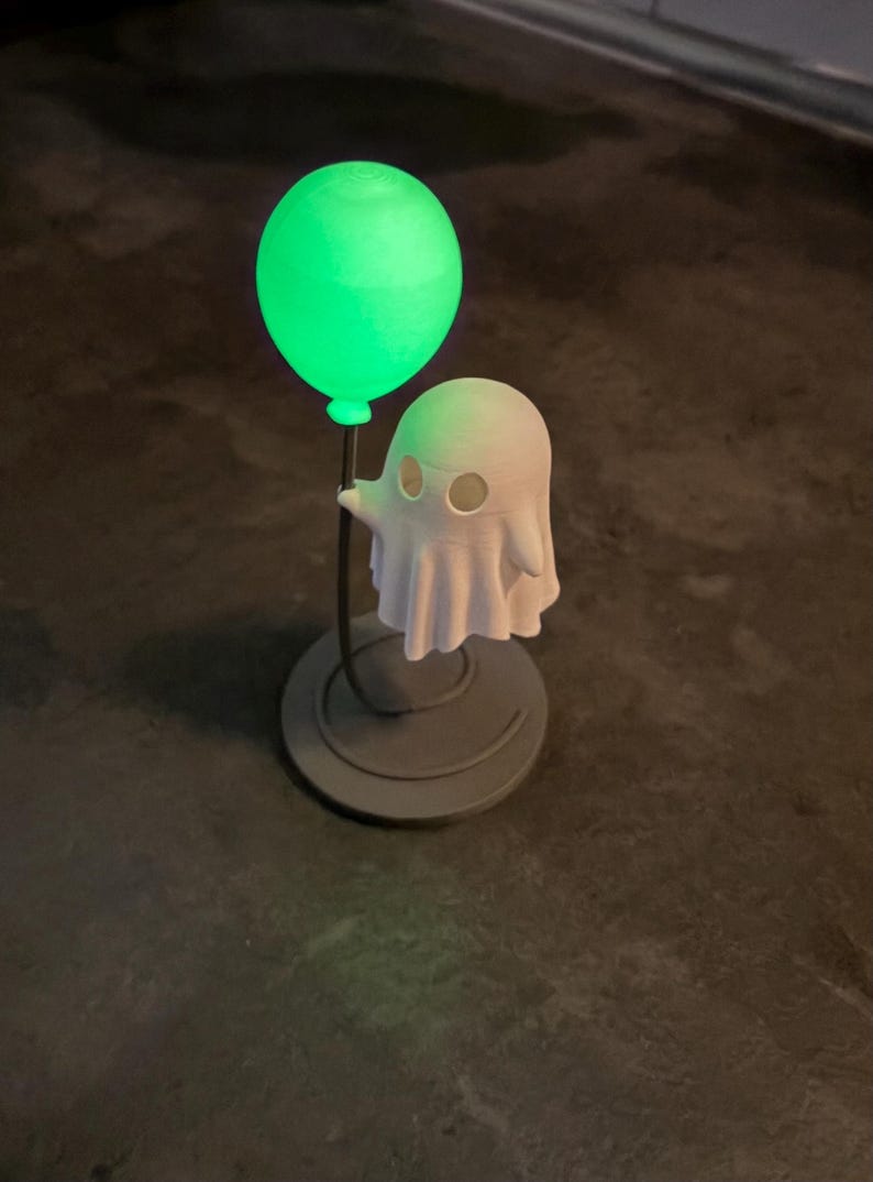 May include: A Halloween-themed ornament depicting a white ghost figure holding a glowing green balloon. The ghost has eye holes and a draped sheet design. The balloon and ghost are mounted on a dark grey circular base.