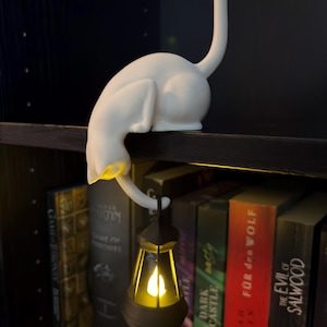 May include: A white cat figurine with an arched back and an upright tail, perched on a shelf edge. The cat holds a small, dark-colored lantern in its front paw, which emits a warm, glowing yellow light from within its translucent structure.