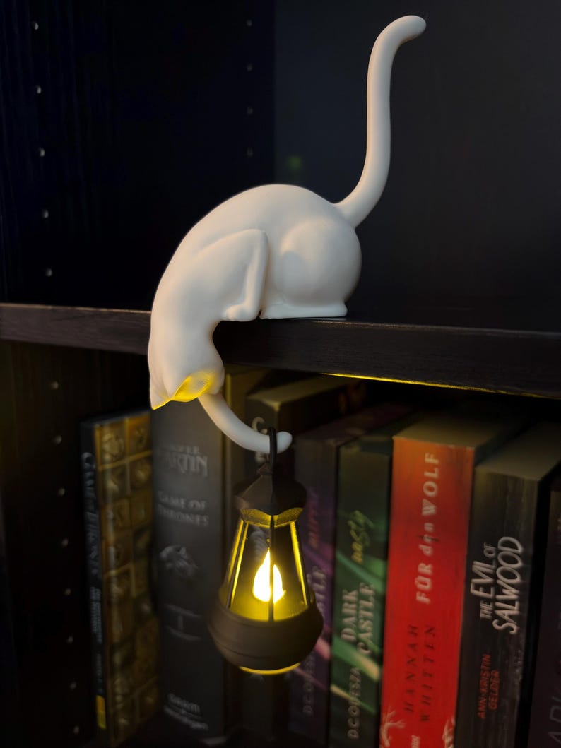 May include: A white cat figurine with an arched back and an upright tail, perched on a shelf edge. The cat holds a small, dark-colored lantern in its front paw, which emits a warm, glowing yellow light from within its translucent structure.