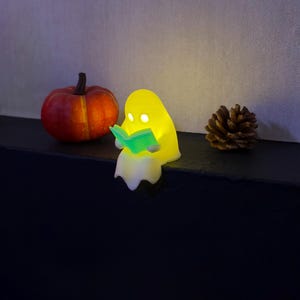 May include: A small, glowing yellow ghost figurine sitting on an edge, holding an illuminated light-green open book. The ghost has two circular cutouts for eyes and a smooth, rounded shape. Beside it sits a small orange pumpkin and a pinecone.