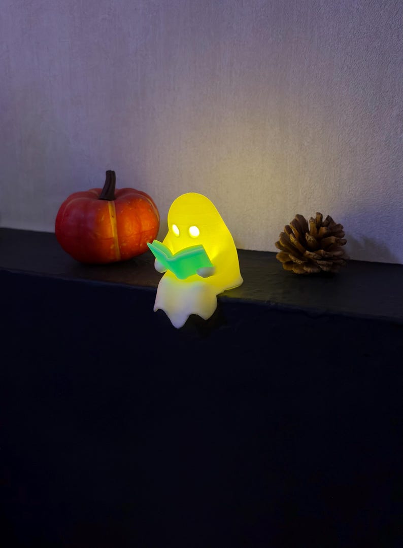 May include: A small, glowing yellow ghost figurine sitting on an edge, holding an illuminated light-green open book. The ghost has two circular cutouts for eyes and a smooth, rounded shape. Beside it sits a small orange pumpkin and a pinecone.