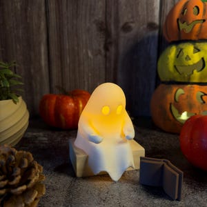 May include: A small, white, ghost-shaped lamp with glowing yellow eyes sits on a light-colored wooden block. The ghost figure is positioned next to a small, open, dark-colored book, with various pumpkins and a pinecone nearby.