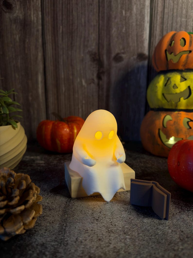May include: A small, white, ghost-shaped lamp with glowing yellow eyes sits on a light-colored wooden block. The ghost figure is positioned next to a small, open, dark-colored book, with various pumpkins and a pinecone nearby.